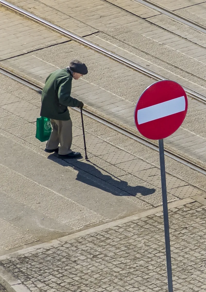 Elderly man with a cane walking past tram tracks toward a red no-entry sign seen from above